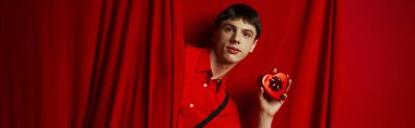 young man in red shirt with suspenders holding heart shaped gift near curtain, 14 February banner clipart