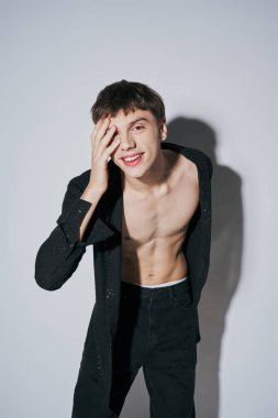 Siyah tişörtlü ve kot pantolonlu kaygısız genç adam gri arka planda eli yüzü ile poz veriyor.