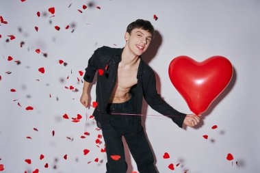 cheerful man in jeans and shirt holding red balloon near heart shaped confetti on grey background clipart