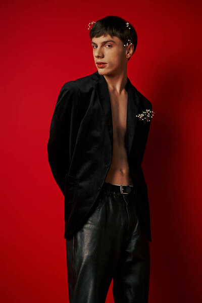 красивый мужчина в стильном наряде с цветами в волосах, смотрящий в камеру и позирующий на красном фоне