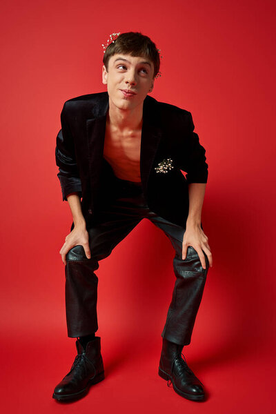 joyful man in stylish outfit with flowers in hair and pocket of velvet blazer looking away on red