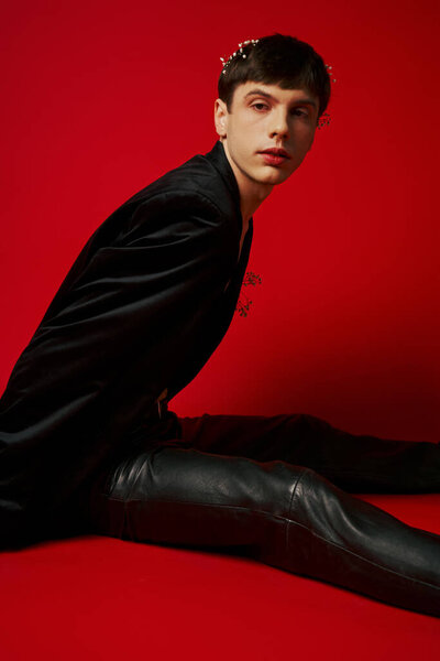 красивый молодой человек в черной одежде с цветущими цветами в волосах сидя на красном фоне