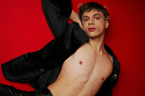 top view of male model in jacket and flowers in hair lying and looking at camera on red background
