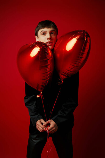 handsome young gentleman in black attire holding heart-shaped balloons on red background