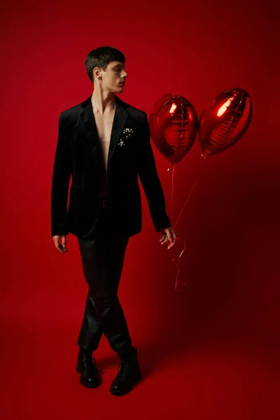 stock image full length of handsome gentleman in black attire holding heart-shaped balloons on red backdrop