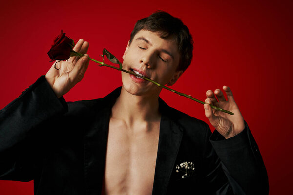 без рубашки молодой человек с закрытыми глазами, позируя в бархатной куртке держа розы в зубах на красном фоне