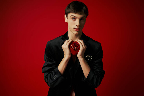 romantic guy holding heart-shaped present and looking at camera on red background, Valentines day
