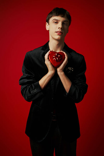 romantic man holding heart-shaped present and looking at camera on red background, Valentines day