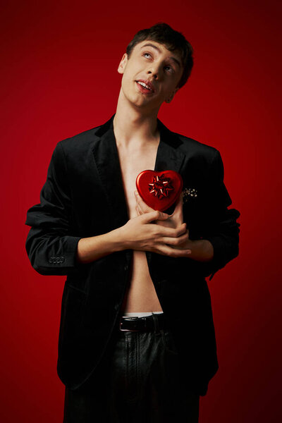 dreamy romantic man in velvet blazer holding heart-shaped present on red background, Valentines day