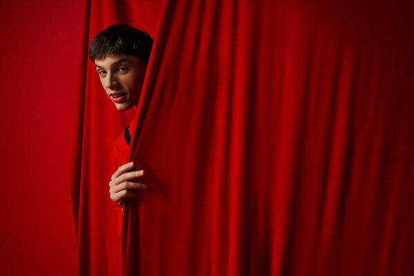 playful young man in vibrant shirt hiding behind red curtain while playing hide and seek, peeking