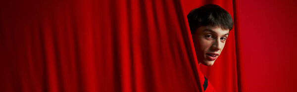 playful young man in vibrant shirt hiding behind red curtain while playing hide and seek, banner