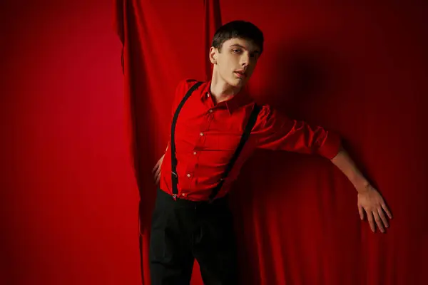 Stock image young man in vibrant shirt and shorts with suspenders standing near red curtain, trendy look