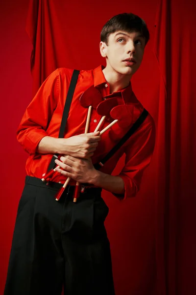 stock image young cupid guy in shirt and suspenders holding heart shaped arrows on red background, 14 February