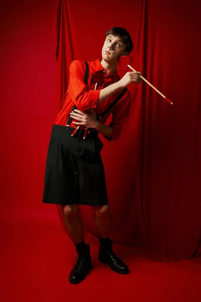 Stock image cupid in shirt and suspenders holding heart shaped arrow near cheek on red background, 14 February