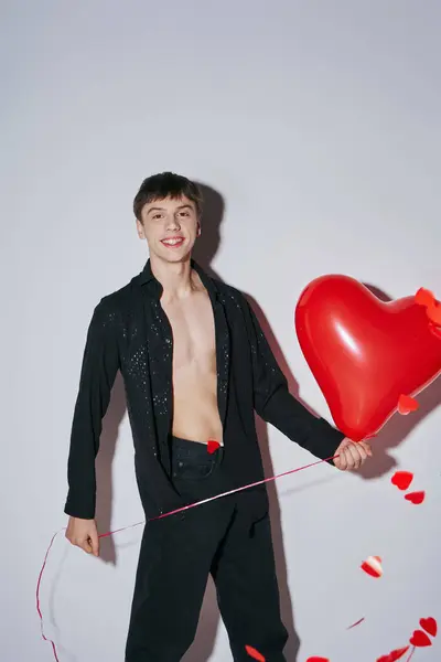 stock image young man in jeans and open shirt holding red balloon near heart shaped confetti on grey background