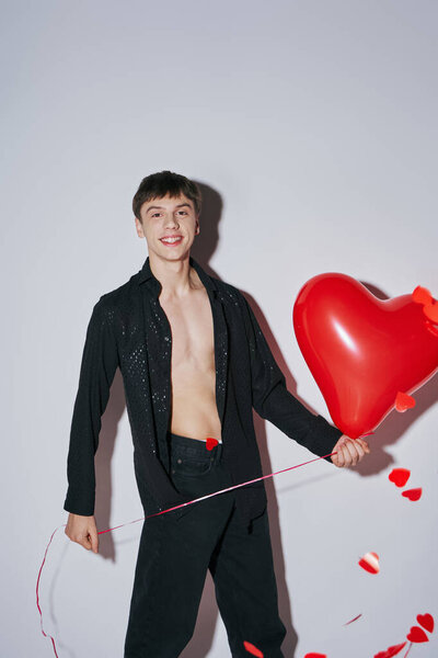 young man in jeans and open shirt holding red balloon near heart shaped confetti on grey background
