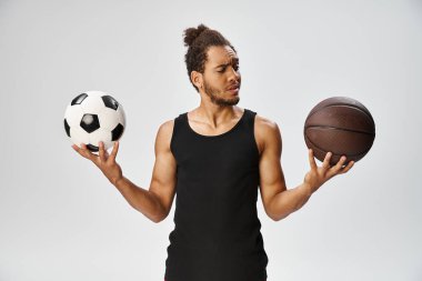 Kafası karışmış Afro-Amerikan sporcusu elinde futbol ve basketbolla gri zeminde poz veriyor.
