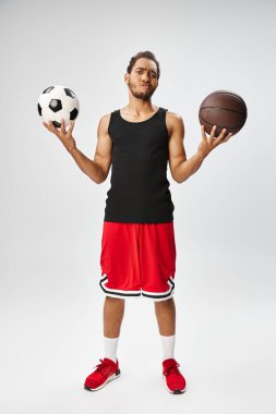 Kafası karışmış Afro-Amerikan sporcusu elinde basketbol ve futbolla gri arka planda poz veriyor.