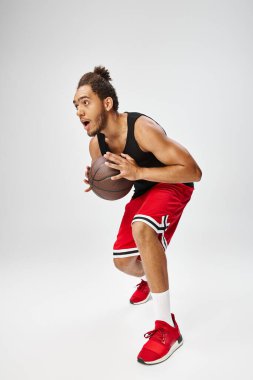 Spor giyimli Afro-Amerikan genç adam gri arka planda basketbol oynuyor.