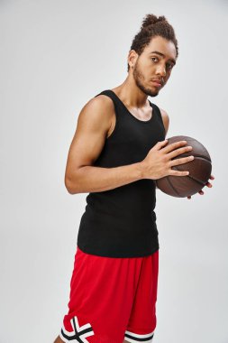 Rahat spor giyimli yakışıklı Afro-Amerikan bir adam basketbol topu tutuyor ve kameraya bakıyor.