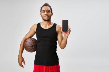 Basketbol ve telefon tutan neşeli Afrikalı Amerikalı adam kameraya bakıyor, online bahis oynuyor.