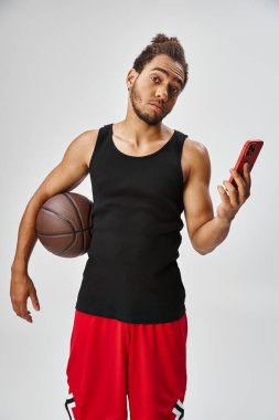 Basketbol ve telefon tutan yakışıklı Afro-Amerikalı adam kameraya bakıyor, online bahis oynuyor.