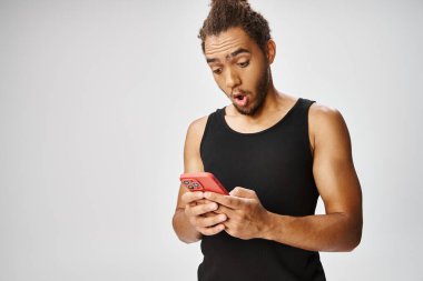 Atletik Afrikalı Amerikalı adamın gri arka planda akıllı telefonuna bakmasına şaşırdım.