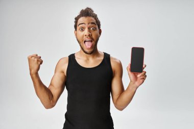 Spor giyimli neşeli Afrikalı Amerikalı adam elinde akıllı bir telefonla kameraya bakıyor, internette bahis oynuyor.