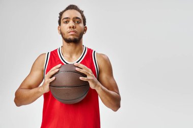İyi görünümlü Afro-Amerikan spor elbiseli adam basketbol topunu tutuyor ve doğrudan kameraya bakıyor.