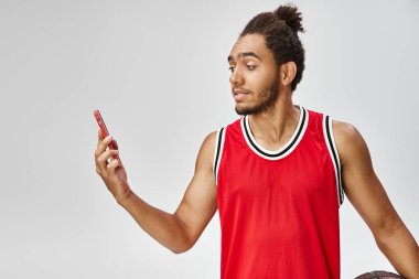 Yakışıklı Afro-Amerikalı adam basketbol topunu tutuyor ve telefonuna bakıyor, internette bahis oynuyor.