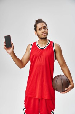 Kafası karışmış Afro-Amerikan bir adam telefonla basketbol oynuyor ve kameraya bakıyor, online bahis oynuyor.