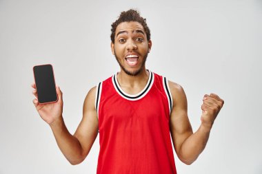 Spor giyimli neşeli Afro-Amerikan adam elinde telefonla kameraya bakıyor, internette bahis oynuyor.