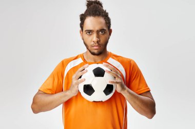 Spor giyimli yakışıklı Afro-Amerikan bir adam futbol topu tutuyor ve kameraya bakıyor.