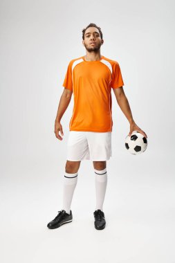 Gri arka planda futbolla poz veren Afro-Amerikan spor giyimli yakışıklı genç bir adam.