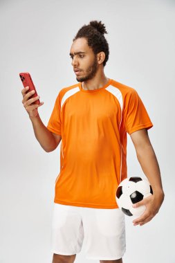 Yakışıklı atletik Afrikalı Amerikalı adam elinde futbol topuyla telefonuna bakıyor, internette bahis oynuyor.