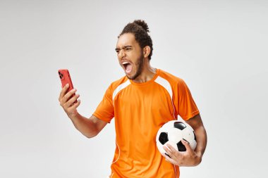 Duygusal Afrikalı Amerikalı erkek spor giyimli, elinde futbol topu, telefona bakıyor, internette bahis oynuyor.