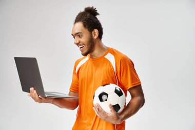 Spor giyimli neşeli Afro-Amerikan adam elinde futbol topuyla dizüstü bilgisayara bakıyor, internette bahis oynuyor.