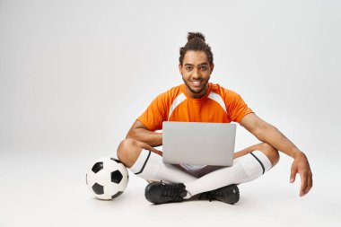 Neşeli Afrikalı Amerikalı adam yerde top ve dizüstü bilgisayarla oturmuş kameraya bakıyor, internette bahis oynuyor.