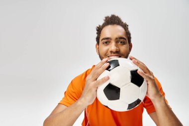 Neşeli Afro-Amerikan adam futbolla poz veriyor ve gri arka planda kameraya bakıyor.