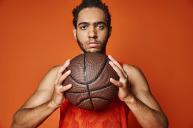 Genç, Afro-Amerikan sporcu, elinde basketbol topuyla turuncu arka planda kameraya bakıyor.