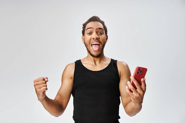 joyous sporty african american man holding mobile phone and looking at camera, online betting
