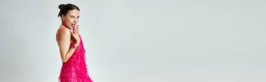 Pembe elbiseli güzel utangaç kız, gri arka planda dudaklarını kapatarak şakacı bir şekilde gülümsüyor.