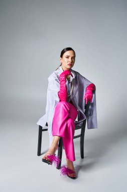 Pembe eldivenler, tüylü ayakkabılar ve şık pantolonlar giyen kendine güvenen esmer kız, sandalyede oturuyor.