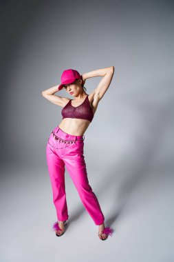 Kırmızı bluzlu, gri arka planda pembe pantolonlu ve şapkalı, elleri başının üstünde bir kadının tam boy fotoğrafı.