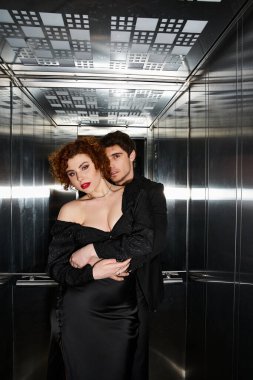 Seksi, çekici, siyah elbiseli ve takım elbiseli çift asansörde sarılıyor ve kameraya bakıyor.