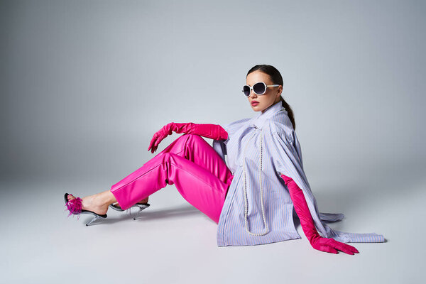 Красивая женщина в розовых перчатках, кожаных штанах и шикарных солнечных очках сидит на сером фоне