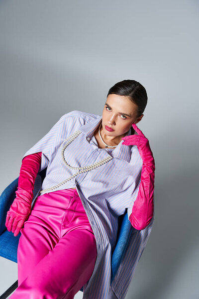 Стильная брюнетка в розовом наряде прикасается к лицу, сидя на стуле на сером фоне