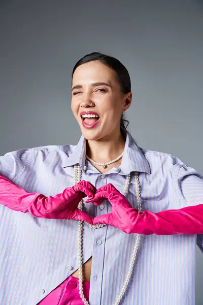 Smiling winking brunette woman with pink trendy gloves making hand heart isolated on grey background