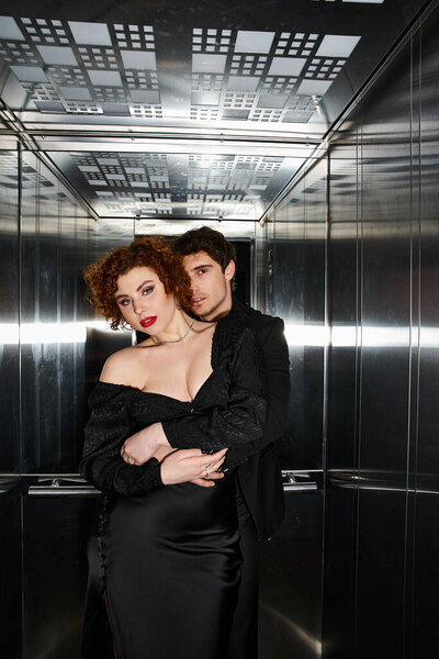 sexy appealing couple in chic black dress and suit hugging in elevator and looking at camera