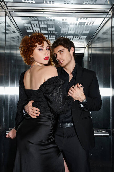 sexy enticing couple in chic black dress and suit hugging in elevator and looking at camera